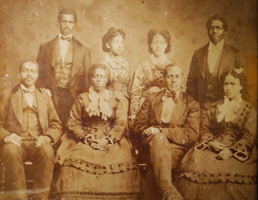Eight African American people pose for a photo: four men and four women. They are in two rows, two women stand between two men in the back and the front row alternates. They are dressed in suits and high quality dresses. They're the storer college singers