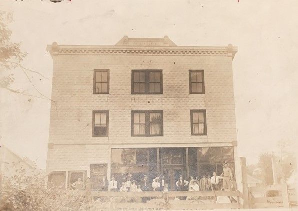 A picture of a large building with three stories, six windows on the front and a large porch wrapping around the ground level. The picture has a faded, white hue.