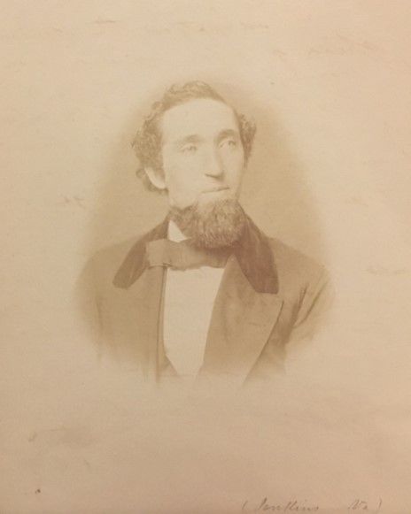 A cream and sepia low contrast image of a man with a beard, wavy hair, a jacket and a necktie. He looks off to the side thoughtfully.