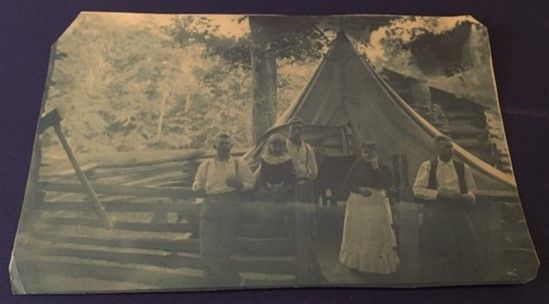 A faded blue tinted image of people sitting outside, facing the camera and backs to a tent. they stand against a log fence.