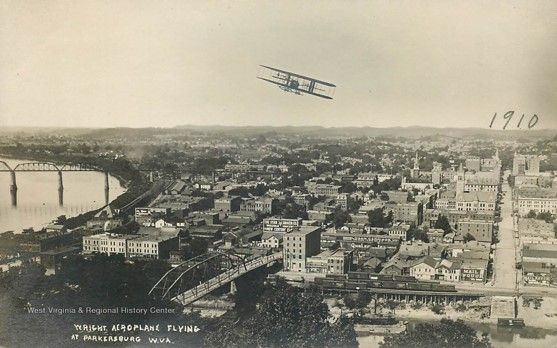 An aerial view of Parkersburg, a city next to a river, you can see hills far off in the distance. Overhead, an airplane flies. 