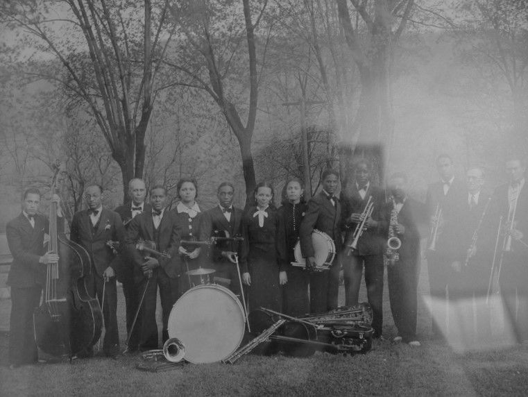 A group of individuals stands outside, holding instruments. A large drum sits in front of them. 