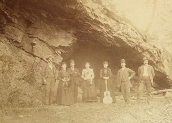 A group of 7 men and women standing at the triangular mouth of a cave, or rock formation. One woman holds an acoustic guitar.