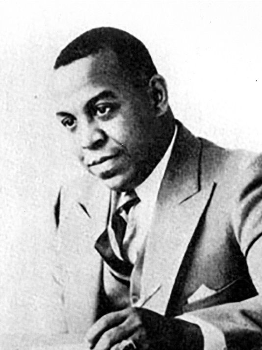 An African American man in a suit and striped tie holds a pencil in his hand, which rests on the table in front of him. He looks thoughtfully to the side, away from the camera. 