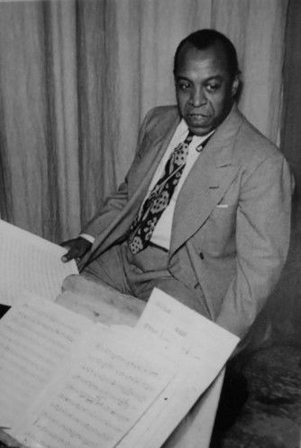 A middle-aged African American man in a suit and tie sits in front of a table covered in sheet music. He looks to the side, past the table. His expression is serious and thoughtful. 