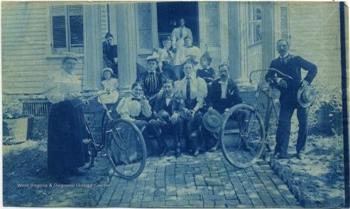 A blue tinted image of several people standing outside the front of a large house. Two are holding bicycles.
