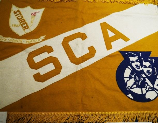 Large dark gold cloth banner with a diagonal stripe of white up the middle reading "SCA." On the upper left corner is a Storer college shield logo and in the lower right is a blue logo of two men surrounded by a circle