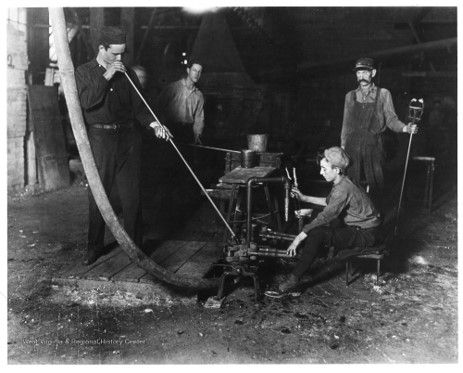 Four people, 3 men and a boy, sit as one blows glass and the boy helps mold the glass.