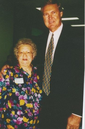 Jerry West, middle-aged poses with ann. He wears a suit and has his arm around her. She has glasses and a black shirt with bright yellow, green and magenta flowers on it. 