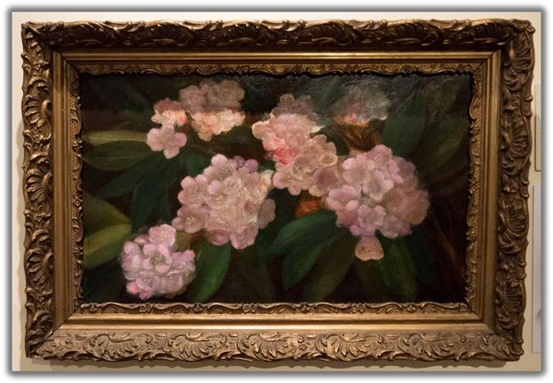 a painting of light pink rhododendrons against a backdrop of dark green leaves. There is a cold embellished frame surrounding the painting.