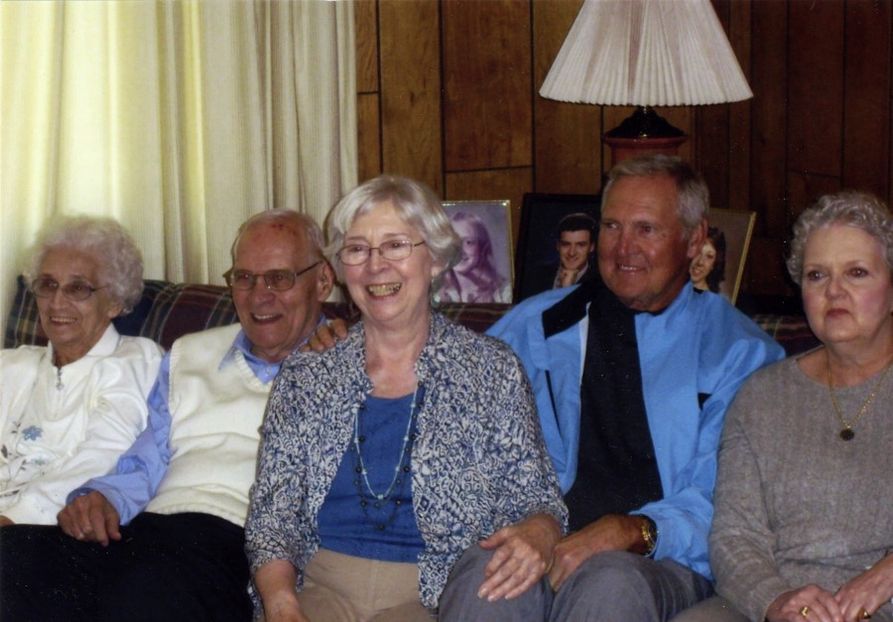 Five elderly people sit on a couch, three women and two men. They are all wearing blue, gray, or white. They smile at the camera