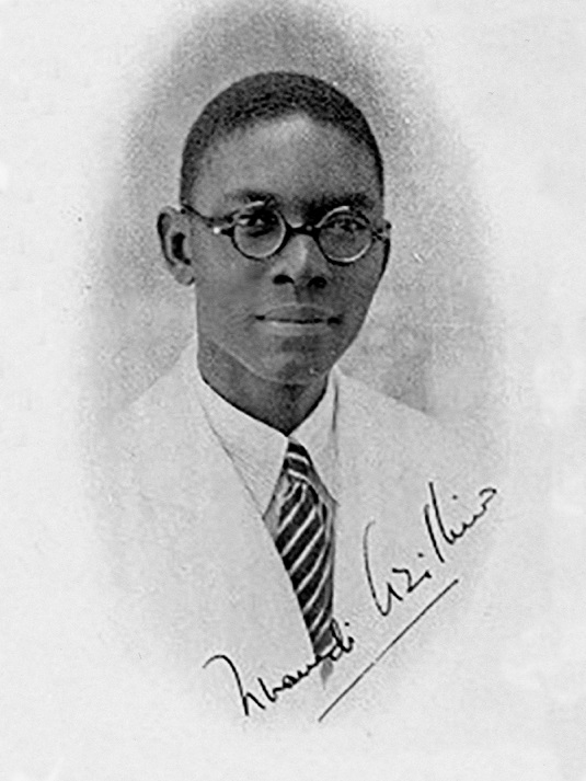  A young African American man with round rimmed glasses looks mildly into the camera. He is wearing a light colored suit and tie.