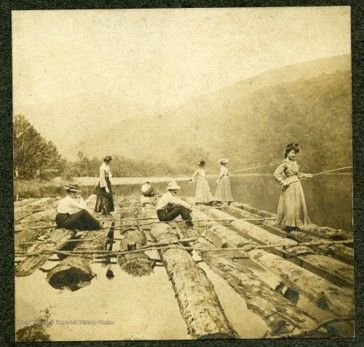 A group of several people in light-colored clothes standing on a dock made of logs overlooking the Cheat River. A woman holds a fishing rod over the edge of the dock
