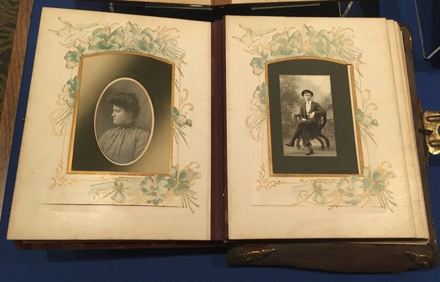 A photo album with an oval portrait of a woman on the left page and a  man on the right. the images have golden accents around the frames, and on the paper behind are detailed illustrations of flowers and greenery. 