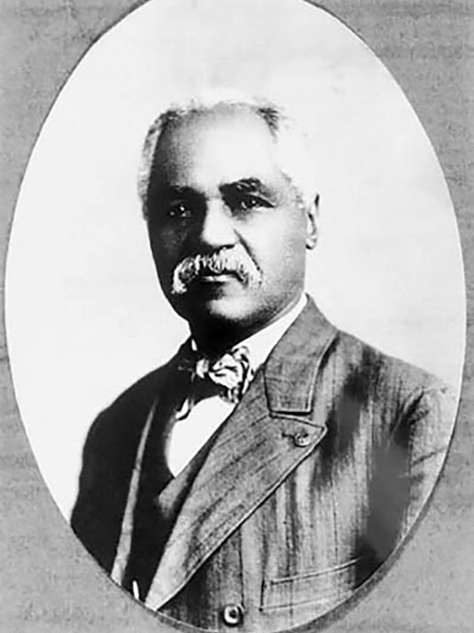 A middle-aged African American man with white hair, and a mustache. He wears a suit and bowtie and looks slightly out of frame. 