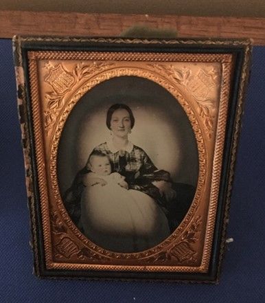 An oval shaped, ornately framed photo of a woman in a plaid dress with a white collar holding a baby wearing a long white smock