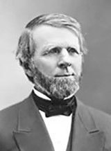 A middle-aged man with a gray beard and medium length hair looks to his left. He is wearing a jacket, a stiff high-collared shirt and a necktie 