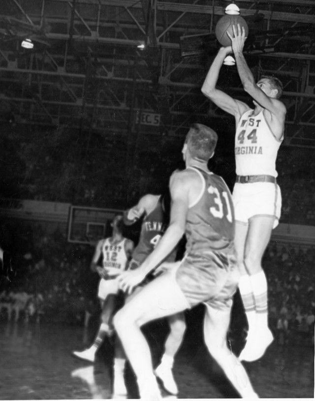 Black and white photo, four players in motion. Jerry West is mid-air, jumping straight up with the ball in his hands, preparing to make a jump shot