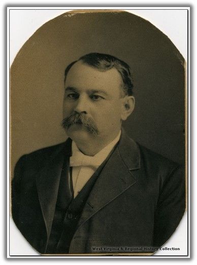 a middle aged man with oiled hair and a large mustache looks past the camera. he is wearing a jacket over a vest and white shirt and bowtie