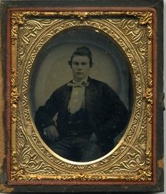 A small ornately framed photo of a man with styled dark hair, a dark suit jacket and vest, a white shirt and bowtie. He looks into the camera. 