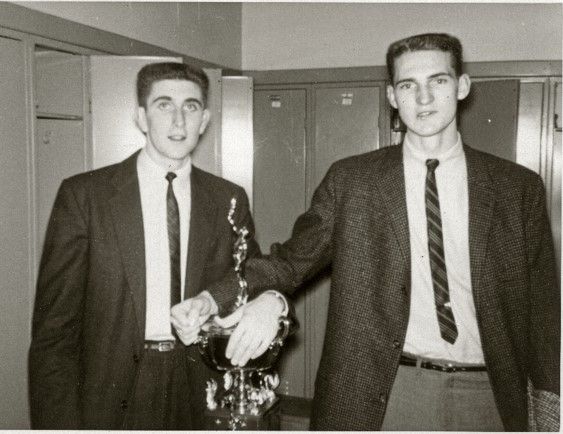 Two young men stand, wearing suit jackets and ties. They both have the same flat top hair cut and look into the camera. They each have a hand on a trophy that sits in front of them 