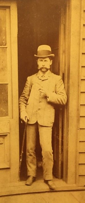 A gold-toned picture of a man in a suit, bowler hat, and carrying a cane. He has a thick mustache and is standing in a doorway