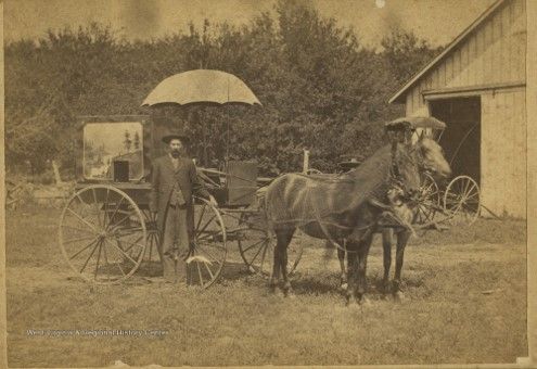 A man in a suit stands in front of a small wooden cart with large wheels and an umbrella over top, pulled by a horse.