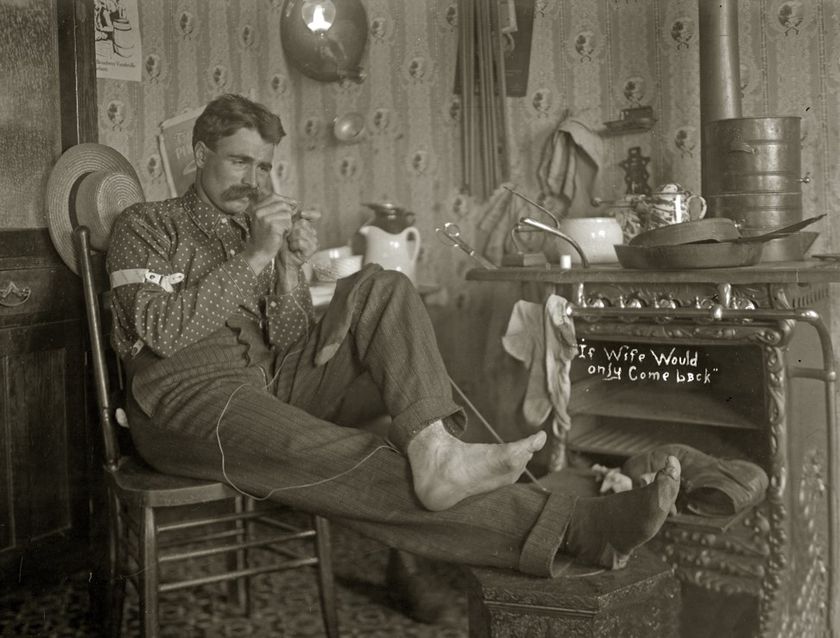 Photo of a man sitting in a wooden chair with his feet up, next to an old fashioned stove with pans and a kettle on top, he looks with focus at a needle and thread in his hands