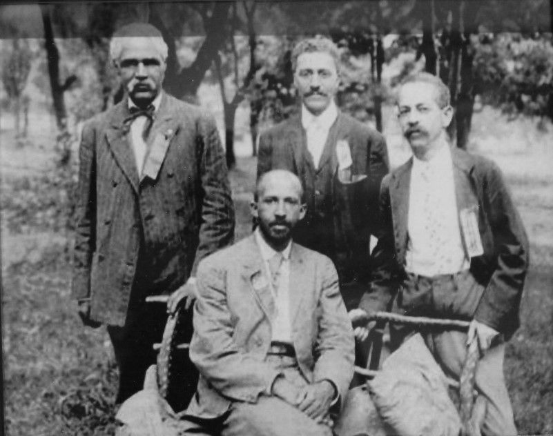 Four men outside on the grass. Three stand in a line and the fourth sits in a chair in front of them. They are wearing suits and white shirts