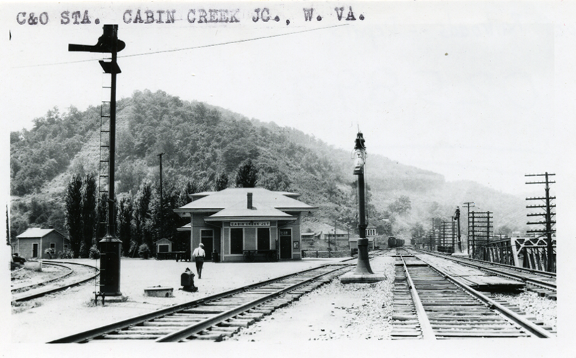 A railroad depot sits in front of a large mountain covered with trees. Multiple railroad tracks lead past it; between the tracks are lamp posts. The ground is covered with a light layer of snow