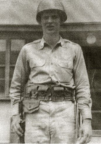A young man wearing a light colored jumpsuit, a leather belt with several pouches, and a hard military issue helmet. He looks seriously into the camera.