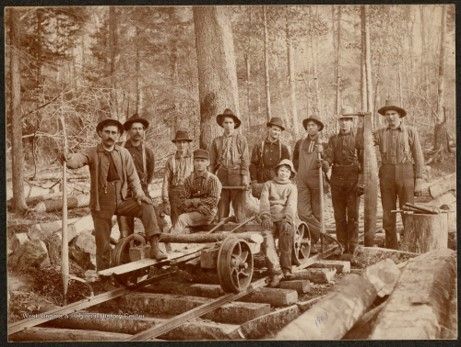 A group of men in work wear stand around a mine cart on a track outside in the woods