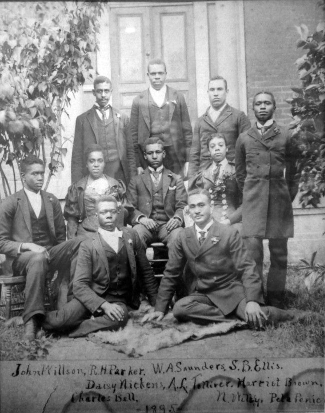 A group of ten African American individuals, dressed in suits and elegant gowns. Four men make up the back row, two men and two women sit in the middle, and two men sit on the ground in the front. They are sitting on a blanket on the grass outside.