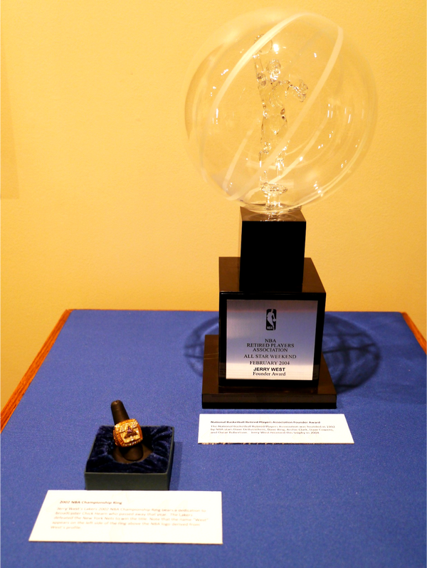 A 2002 NBA gold championship ring. To the right is an award, a large orb made of clear glass, inside is a clear statue of a player. it is on top of a two tier dark wooden base