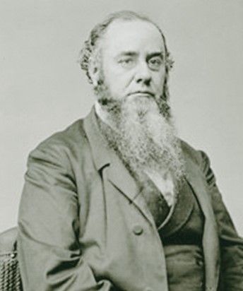A middle-aged man with graying hair and a beard down to his chest looks to his left away from the camera. He is wearing a vest and jacket, and has a serious expression on his face
