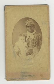 A black woman wearing a plaid dress and white neckerchief and head wrap holds a baby on her lap. The baby is wearing a white gown.