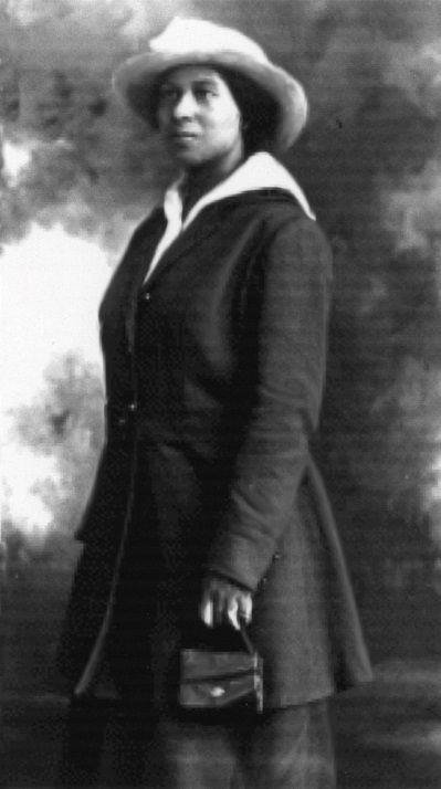 A black woman wearing a white hat and a dark overcoat over a skirt, looks to the left. She is holding a small purse in her hand. 