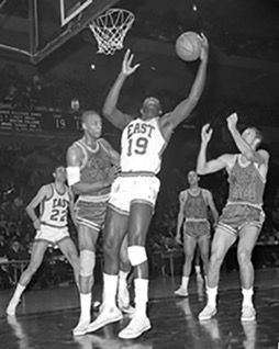 A black and white image from the all-star game. 5 players are seen, two wearing white East uniforms, and three wearing darker West unifroms. the man in the middle, who is african american, lunges upward, as the players surrounding him reach forward and up