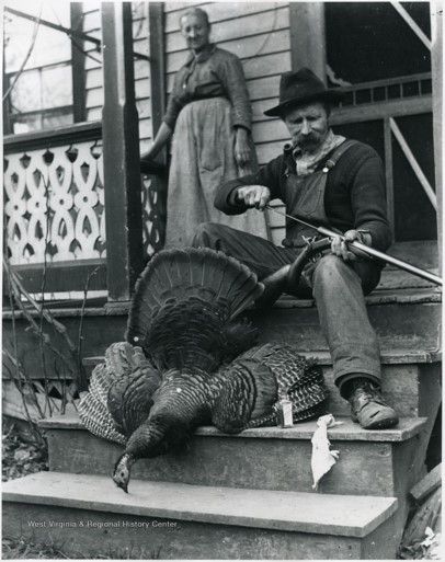 A man sits on the stoop of a house holding a hunting rifle with a large turkey laying next to him. Behind him, a woman stands against the wall and looks down at him