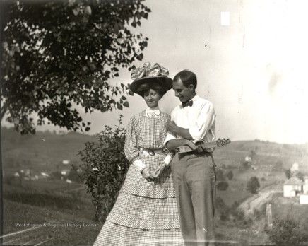 A young woman in a layered checkered dress and large hat looks down and stands next to a young man in a white shirt. He looks down at her smiling and one of his hands are against her shoulder