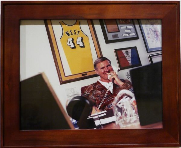Jerry sits in his laker's office. he is using a white landline phone and smiling. Behind him, several lakers-related items and awards, including his jersey, are displayed on the wall 