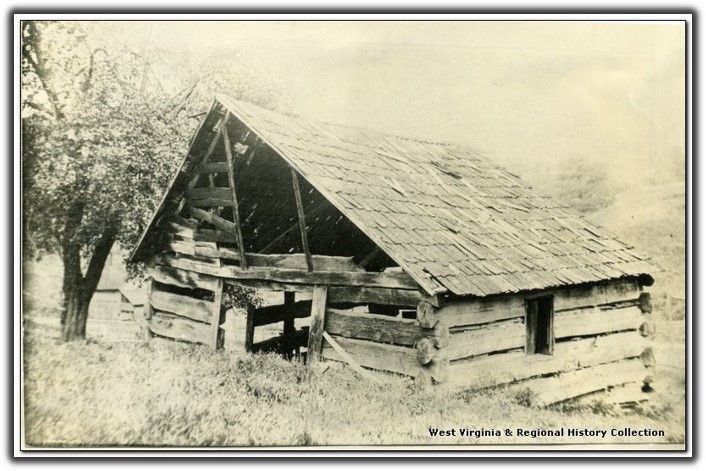 A faded black and white image of a wooden schoolhouse with a simple roof. It looks to be one room.