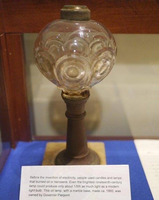 An oil lamp from 1860. It has a narrow, short base and a round globe on top with circular scale-like designs. 