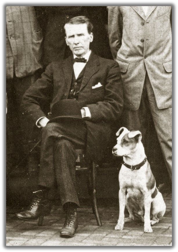 a man in a suit and bowtie sits in a wooden chair. to his left is a white and spotted dog with a collar