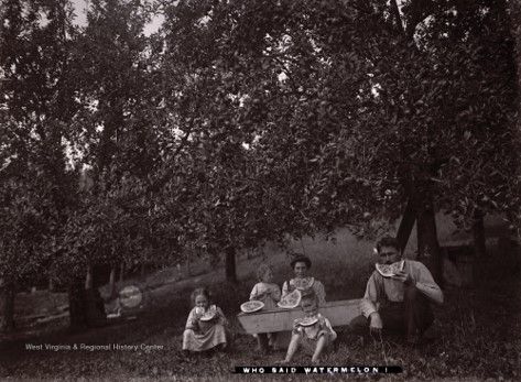 A family sits at a wooden table in a field surrounded by trees, eating watermelon 