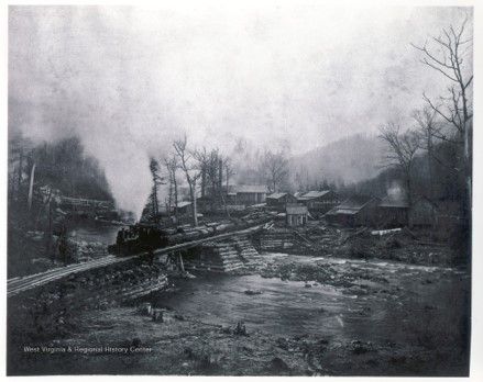 A train carrying log carts travels across a small bridge, surrounded by trees and mountains