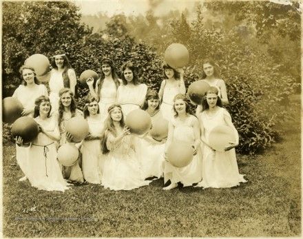A group of girls pose in a grassy field surrounded by hedges. They wear white dresses and hold exercise balls in their hands. 
