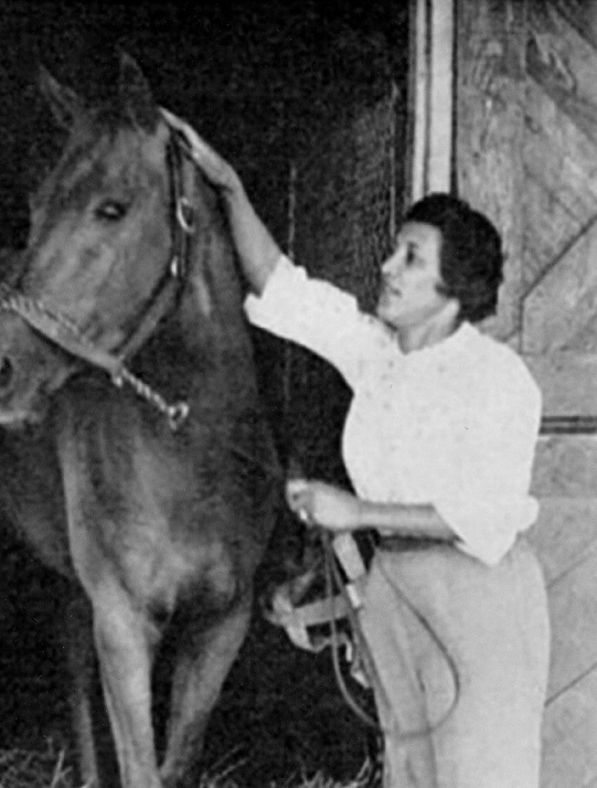 A woman in a white shirt and light colored trousers stands next to a horse. In one hand she holds the reigns, with the other she pets behind the horse's ears. She is smiling 