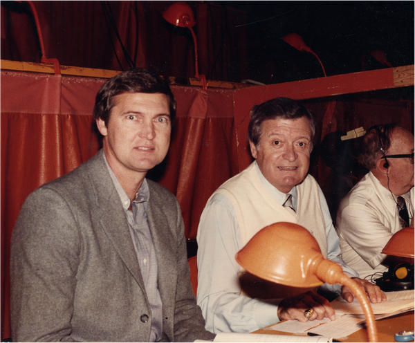 Jerry wearing a light gray suit jacked and button down, sitting next to a middle aged man in a white weater, tie and button down. they both look into the camera