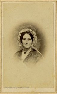 A portrait of a woman on a cream background. She wears a dress, a bonnet, and has her dark hair parted down the middle. She looks straight into the camera. 